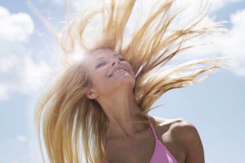 Una donna bionda con un costume da bagno rosa, sorride con i capelli che volano al vento e il sole che splende.
