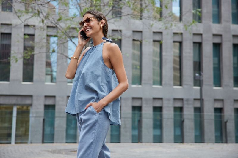Donna che parla al cellulare indossa un abito di lino e occhiali da sole mentre cammina all'aperto vicino a un edificio moderno della città