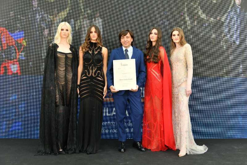 Gruppo di persone con modelle in abiti eleganti, uomini in giavva e cravatta e uomo che tiene targa di riconoscimento in mano