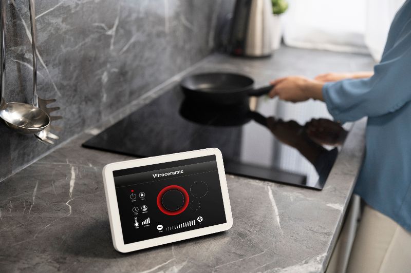 Primo piano di un tablet bianco su un ripiano grigio in una cucina moderna, che mostra un'interfaccia di controllo per un piano cottura a induzione, con una persona che usa una padella sullo sfondo.