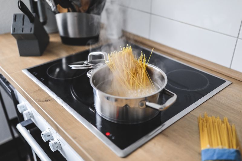 Una pentola di spaghetti in ebollizione su un moderno piano cottura a induzione nero, con una confezione di pasta cruda e un ceppo di coltelli in legno sullo sfondo di una cucina.