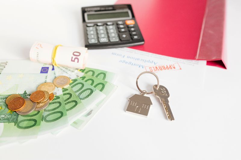 Banconote da 100 euro, monete, un rotolo di banconote da 50 euro, chiavi di casa con portachiavi a forma di casa, una calcolatrice nera e una cartella rossa "APPROVED" su una superficie bianca.