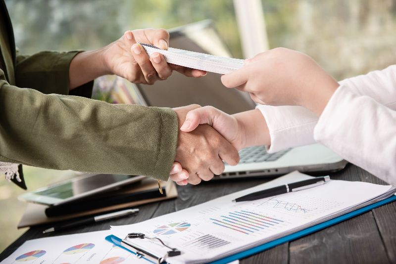 Due persone si stringono la mano sopra una pila di banconote che vengono passate, con documenti finanziari, un laptop e una penna su un tavolo scuro.