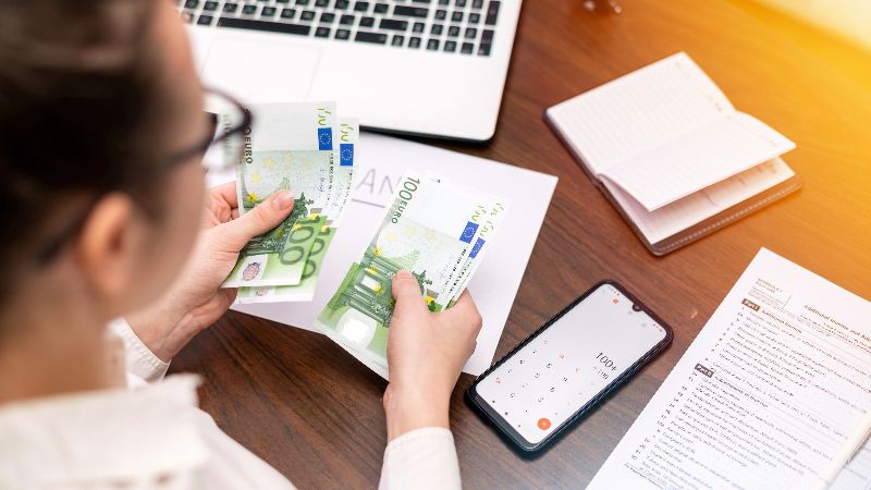 Una persona con occhiali conta una pila di banconote da 100 euro su un tavolo di legno, con un laptop, uno smartphone con calcolatrice e documenti finanziari sullo sfondo.