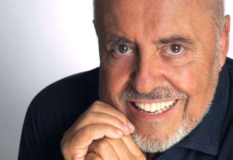 Elio Fiorucci sorride con un'espressione amichevole, mettendo in evidenza la sua barba brizzolata e i suoi occhi scuri, con la mano appoggiata sul mento.
