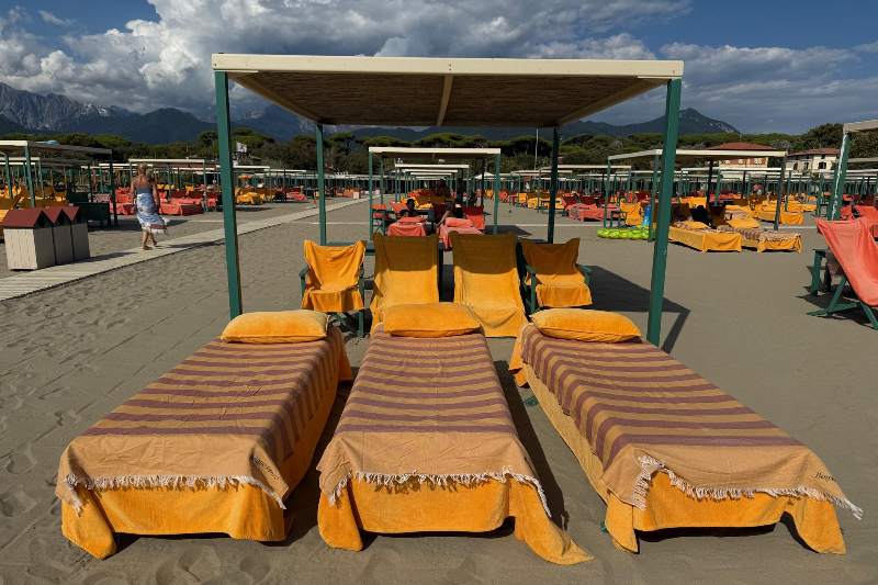 Spiaggia del Bagno Piero a Forte dei Marmi con lettini arancioni a righe gialle e ombrelloni, sotto un cielo parzialmente nuvoloso, in una giornata estiva.