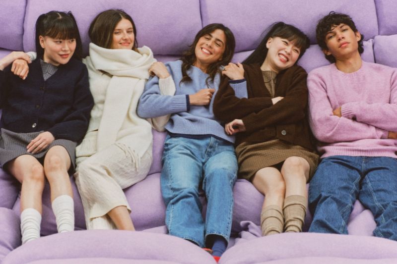 Un gruppo di cinque modelli multietnici sorride, indossando capi in maglia della collezione Uniqlo Soufflé Yarn e seduti su una superficie soffice e viola.