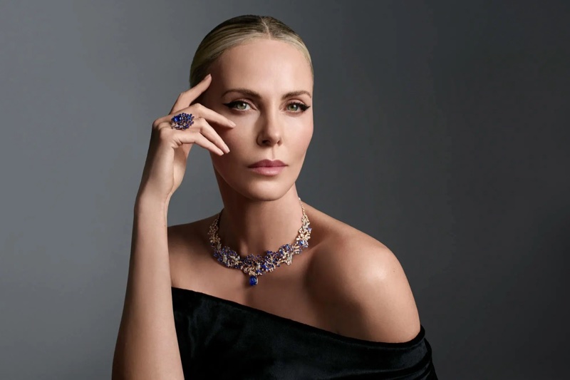 L'attrice Charlize Theron posa con i capelli raccolti, indossa un top nero monospalla e un'elegante parure di gioielli Dior con pietre blu scure e chiare.