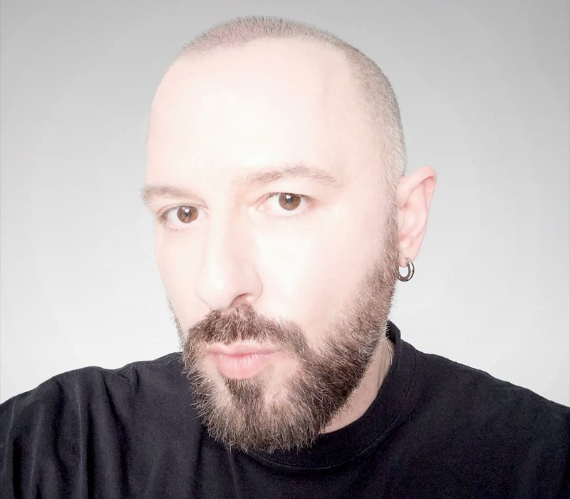 Primo piano di Demna Gvasalia, Direttore Creativo di Gucci, con barba e capelli rasati, indossa una t-shirt nera e un orecchino a cerchio.