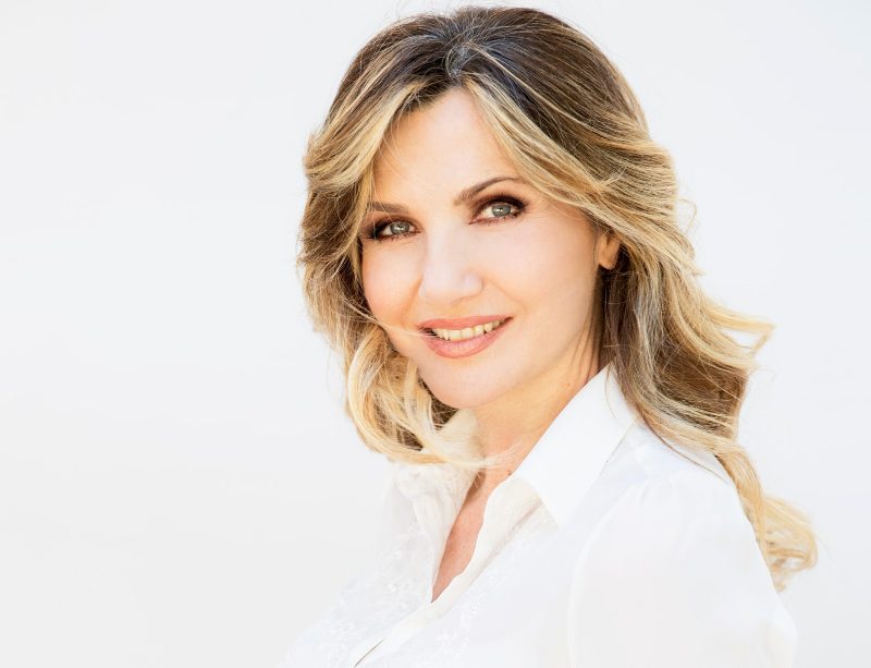 Un primo piano di Lorella Cuccarini che sorride elegantemente, con i capelli biondi sciolti e una camicia bianca.