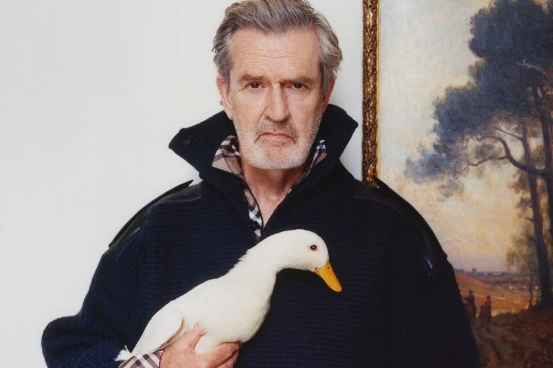 L'attore britannico Rupert Everett posa per la campagna Burberry Inverno 2025 tenendo in braccio un'anatra bianca. Indossa un maglione blu scuro con colletto rialzato che rivela l'iconico motivo Burberry Check