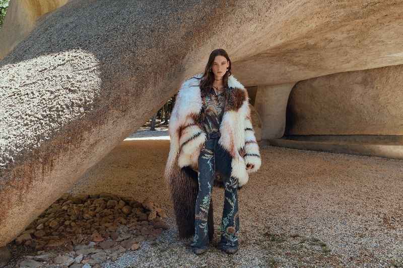 Modella che indossa un cappotto di pelliccia multicolore e jeans a zampa d'elefante con motivi floreali, in posa in un paesaggio roccioso e primordiale nella Campagna Etro Magma FW25/26