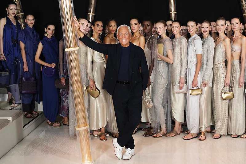 Giorgio Armani con un abito blu scuro e un paio di sneakers bianche, saluta con una mano alzata al centro della passerella, circondato da modelle che indossano abiti lunghi chiari della sua collezione