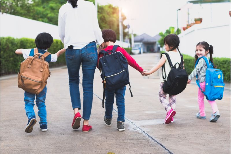 Una donna cammina con diversi bambini che indossano zaini, tenendosi per mano, simboleggiando il ritorno a scuola e la sicurezza
