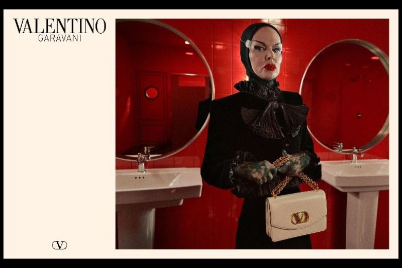 odella con trucco teatrale, cuffia nera e un abito nero con ruches, tiene una borsa Valentino Garavani bianca con catena dorata in un bagno dalle pareti rosse