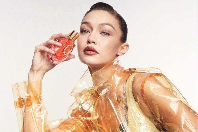 Gigi Hadid posa con il profumo Rabanne Million Gold Pure Diamonds Eau de Parfum indossando un trench trasparente ambrato e un look dorato